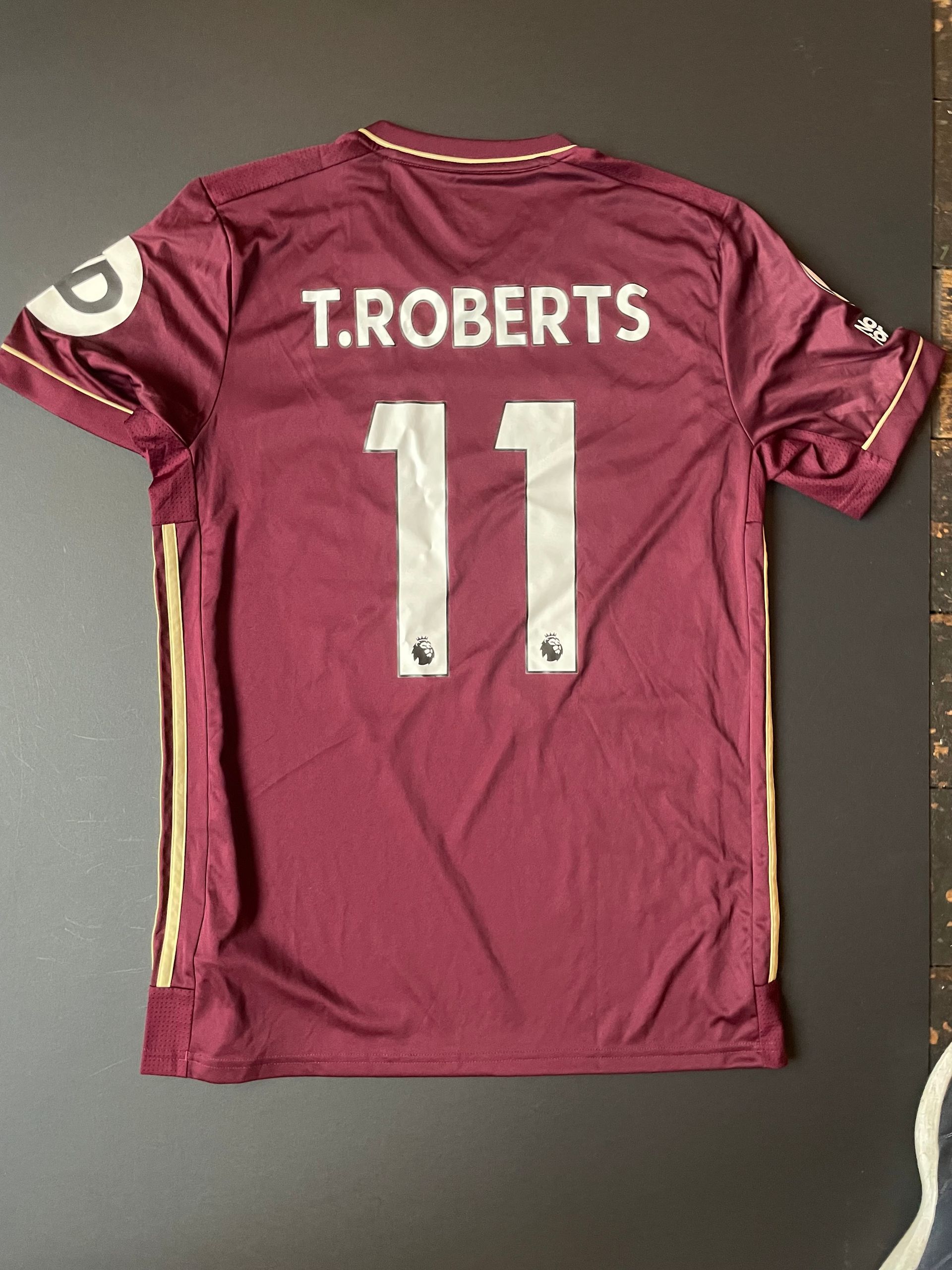Maroon soccer jersey with “T. ROBERTS” and number 11 on the back, laid flat on a dark surface;Maroon soccer jersey with gold stripes and SBOTOP logo laid flat on a dark background
